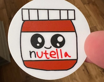 May include: A white circle sticker with a cartoon illustration of a jar of Nutella. The jar has a red lid and a white label with the word "Nutella" written in red. The jar has two black eyes and a small smile.
