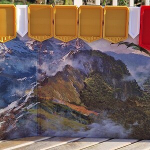 May include: A fantasy game board with a mountain landscape and dragons. The board has red and gold plastic game piece holders. The artwork depicts a detailed mountain range with dragons flying in the sky.
