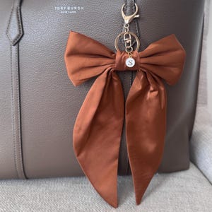 Pode incluir: Um pendente de bolsa em forma de laço de cetim cor de ferrugem com um fecho lagosta e argola de metal dourado. Um pequeno pendente circular branco com a letra S em preto está preso ao centro do laço, que pende sobre uma bolsa de couro cor de toupeira.