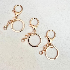 Puede incluir: Tres llaveros de metal en tono dorado, cada uno con una anilla circular unida a un pequeño cierre de mosquetón mediante una cadena corta. Los herrajes tienen un acabado pulido y un diseño consistente, mostrando una estructura metálica funcional.