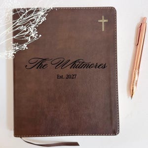 May include: A dark brown leather-like journal with stitched edges, featuring a gold cross icon at the top and the engraved text "The Whitmores" above "Est. 2027" in the center. A rose gold metallic pen is positioned to the right of the journal.