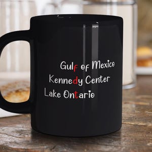 Può includere: Una tazza in ceramica nera con testo bianco che recita Gulf of Mexico, Kennedy Center e Lake Ontario. Le lettere f, d e t sono evidenziate in rosso e piccole icone a forma di cuore bianco sono posizionate accanto al testo sulla tazza.