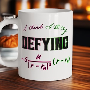 May include: White ceramic mug with the text "I think I'll try DEFYING" in a mix of green, purple, and black fonts. Below the text is a mathematical formula. The mug has a curved handle and is set against a blurred background.