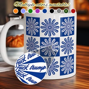 以下が含まれることがあります： 青と白の花柄と「Fuck Trump」の文字が青色のスタイリッシュなフォントで描かれた白いセラミックマグ。マグには白いハンドルがあり、木製の表面に置かれています。