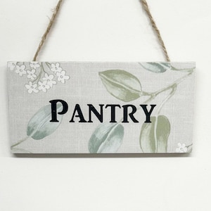 May include: A white wooden sign with a floral pattern and the word "PANTRY" in black lettering. The sign is hanging from a twine string.