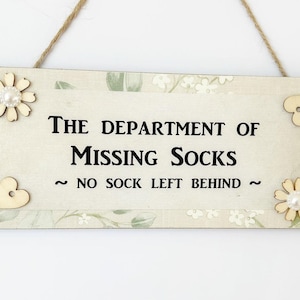 May include: A white wooden sign with green floral accents and the text "The Department of Missing Socks ~ No Sock Left Behind ~". The sign has a white pearl flower and a wooden heart on each side of the text.