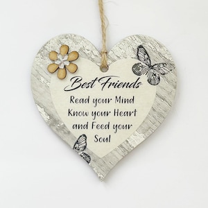 Könnte beinhalten: Ein weißes, herzförmiges Schild aus Holz mit einer abgenutzten Oberfläche. Das Schild hat einen schwarz-weißen Schmetterling, eine gelbe Blume und den Text "Best Friends Read your Mind Know your Heart and Feed your Soul".