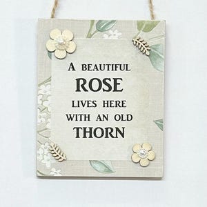 Könnte beinhalten: Ein Holzschild mit floralem Design und dem Text "Eine schöne Rose lebt hier mit einem alten Dorn".