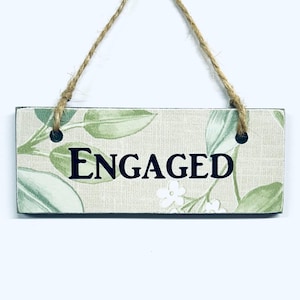 May include: A rectangular wooden sign with the word "ENGAGED" in black capital letters. The sign features a floral design with green leaves and white flowers on a beige background. It is suspended by a natural twine string.