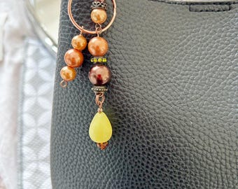 May include: A brown leather handbag with a copper-toned charm attached to the handle. The charm features a ring with brown and gold beads, a yellow teardrop-shaped bead, and a brown bead with green glitter.
