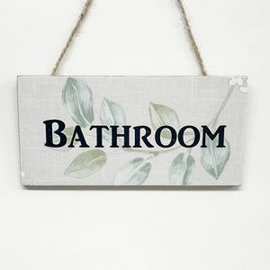 May include: A white wooden sign with a green leafy design and the word "BATHROOM" in black lettering. The sign is hanging from a string.