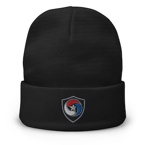 May include: A black cuffed beanie featuring an embroidered shield-shaped patch on the front. The patch displays a circular emblem with three curved segments in red, white, and blue, centered with a small five-pointed star in silver thread.