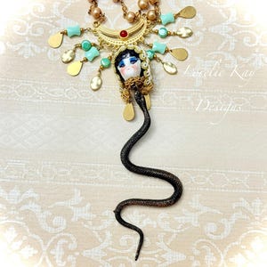 May include: A gold-toned necklace featuring a central porcelain doll head with painted features, framed by a crescent moon charm with a red stone. The piece includes turquoise beads, gold teardrop dangles, and a long, textured black snake pendant.