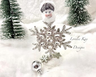 May include: A white porcelain doll head wearing a white fur hat sits atop a sparkly silver snowflake ornament. The snowflake is surrounded by white beads and sits on a white base. The background is a white snowy surface with two small green pine trees.