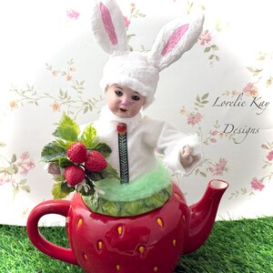May include: A porcelain doll wearing a white bunny costume sits inside a red strawberry teapot. The doll is holding a bouquet of red strawberries. The teapot is sitting on a green grassy surface. The background is a white floral pattern with the text "Lorelei Kay Designs".
