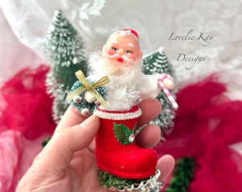 May include: A vintage Santa Claus figurine wearing a white fur coat and red hat, holding a candy cane and a small Christmas tree. He is standing in a red boot with green holly and a white pearl embellishment. The figurine is made of felt and has a whimsical, retro look.