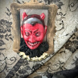以下が含まれることがあります： 悪魔の顔が描かれた額入りの壁掛け。悪魔の顔は赤色で、黒い髪と角、大きな笑顔、黒いフリルの襟が特徴です。フレームは金色で、背景は模様入りの布です。