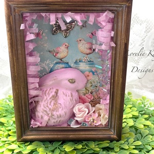 May include: A pink ceramic bunny figurine in a framed shadow box with a blue and white floral background. Two small birds are perched on top of blue and white porcelain vases. The shadow box is decorated with pink tissue paper and a silver butterfly.