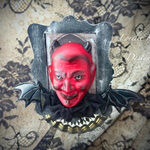 以下が含まれることがあります： 角のある赤い悪魔の顔で、使い古された灰色の長方形に額装され、黒いコウモリの羽で飾られています。悪魔は不吉な笑顔をしており、レースと花の模様の背景に設定されています。「Lorelie Kay Designs」というテキストが表示されます。