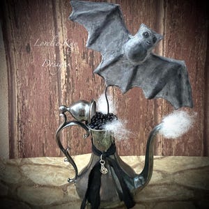 May include: A silver teapot with a black ribbon and a charm hanging from it. A grey paper bat is perched on top of the teapot, which is filled with black beads and white cotton.