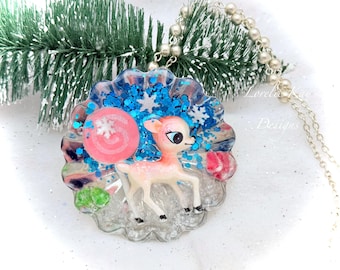 May include: A round pendant necklace featuring a pink deer figurine, a pink lollipop, and white snowflake accents embedded in clear resin with blue glitter. The pendant is attached to a silver-toned ball chain necklace.