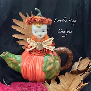 May include: A ceramic teapot shaped like a pumpkin with a green spout and handle. A doll head with blonde hair sits on the lid, adorned with a ruffled orange fabric collar, a tan raffia bow, and a small acorn charm. Text reads Lorelie Kay Designs.