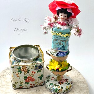 May include: A whimsical tea party centerpiece featuring a porcelain doll wearing a red flower hat, stacked on top of vintage tea tins and teacups decorated with floral patterns. The base is a floral patterned tin with a removable lid.