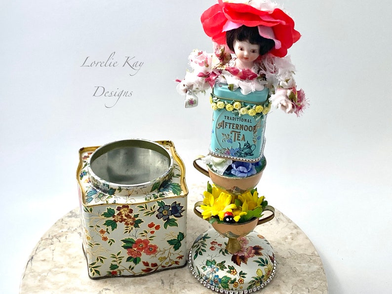 May include: A whimsical tea party centerpiece featuring a porcelain doll wearing a red flower hat, stacked on top of vintage tea tins and teacups decorated with floral patterns. The base is a floral patterned tin with a removable lid.