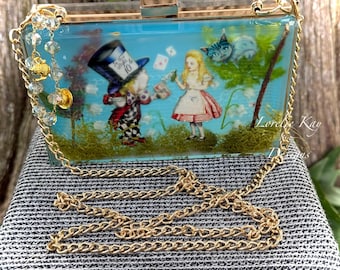 May include: A rectangular clear acrylic clutch bag featuring an Alice in Wonderland scene with the Mad Hatter and Alice. The bag includes a gold-tone metal chain strap, a gold clasp, and decorative crystal beads. The interior contains green moss.