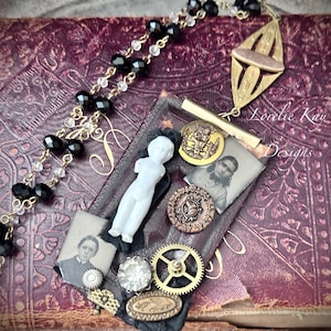 Può includere: Una collana di ispirazione vintage con una catena di perline nere e trasparenti, un ciondolo in ottone con un design a taglio e un'immagine incorniciata con una bambola di porcellana, una fotografia d'epoca e ingranaggi in ottone. Il ciondolo riporta il testo "Lovelie Kay Designs".