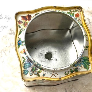 May include: An empty, square, vintage tin with a floral design in shades of blue, green, red, yellow, and orange. The tin has a gold trim and a white background. The inside of the tin is silver.