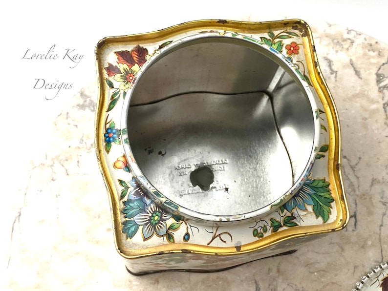 May include: An empty, square, vintage tin with a floral design in shades of blue, green, red, yellow, and orange. The tin has a gold trim and a white background. The inside of the tin is silver.