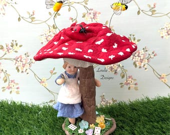 May include: A whimsical figurine featuring a red mushroom with white spots, a small girl in a blue dress and white apron, and two decorative bees. The base includes playing cards and flowers. The text "Lorelie Kay Designs" is visible.