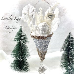May include: A decorative Christmas ornament shaped like a cone, featuring a white cherub head, white floral accents, and a silver and gold glitter design. A silver snowflake hangs from the bottom. Two small green Christmas trees are on either side.