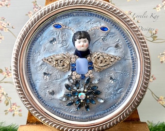 May include: A circular artwork framed in silver, featuring a blue and white butterfly with a porcelain doll head at its centre. The butterfly is adorned with rhinestones and a blue and white cameo. The artwork is signed "Lorelie Kay Designs".