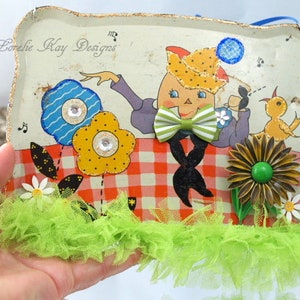 May include: A vintage tin tray with a whimsical illustration of a person wearing a hat and bow tie, sitting on a red and white checkered picnic blanket. The tray is decorated with colorful flowers, a green and yellow bird, and a green and brown flower with a green button center. The tray is surrounded by green tulle.