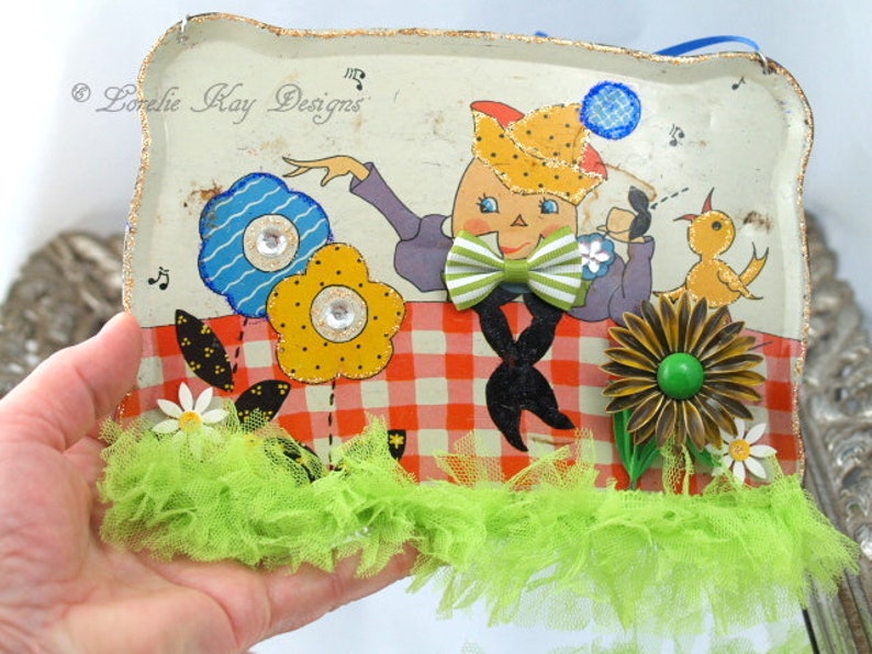 May include: A vintage tin tray with a whimsical illustration of a person wearing a hat and bow tie, sitting on a red and white checkered picnic blanket. The tray is decorated with colorful flowers, a green and yellow bird, and a green and brown flower with a green button center. The tray is surrounded by green tulle.