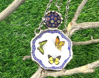 May include: A silver-toned necklace with a pendant featuring three colorful butterflies on a white background with a blue border. The top of the necklace has a round, floral-patterned bead. The background is green grass and a wooden branch.