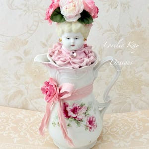 May include: A white porcelain jug decorated with pink floral patterns and a pink ribbon tied with a fabric rose. A doll head with blonde hair, wearing a pink ruffled collar and a crown of pink and white fabric roses, sits inside the jug.