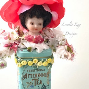 May include: A porcelain doll wearing a red flower hat and a floral dress, standing in a vintage blue tin with the text "TRADITIONAL AFTERNOON TEA".