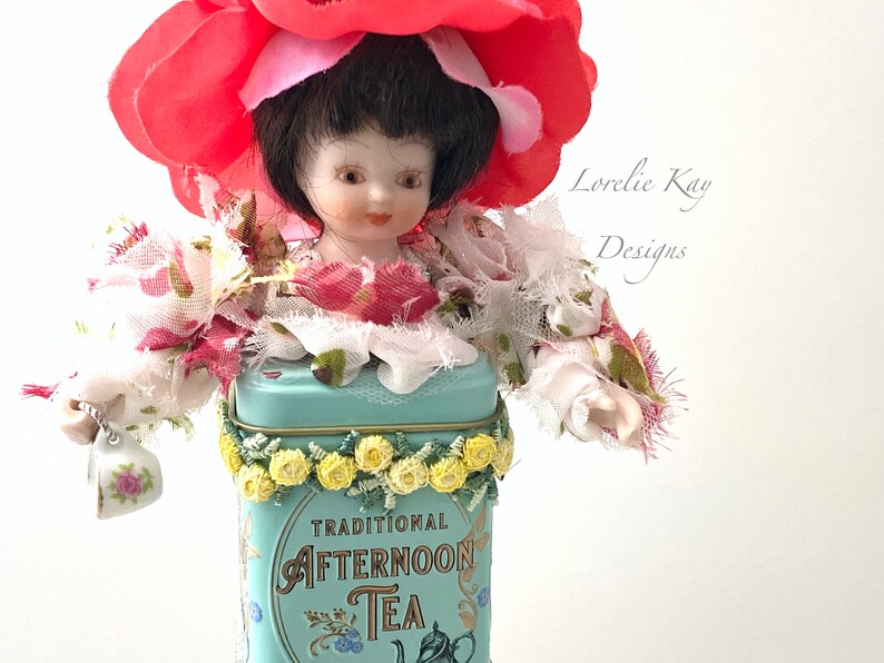 May include: A porcelain doll wearing a red flower hat and a floral dress, standing in a vintage blue tin with the text "TRADITIONAL AFTERNOON TEA".