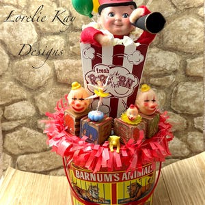 May include: A circus-themed display with a popcorn box holding a clown figure, balloons, and a vintage tin bucket labelled "Barnum's Animals Crackers." The arrangement includes small toy blocks and a ballerina figurine.