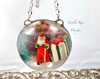 May include: A silver Christmas ornament with a Santa Claus figurine inside. The ornament is hanging from a silver chain and has a white background with a green tree and a red and green gift box.