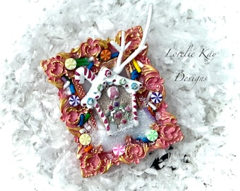 May include: A decorative Christmas ornament with a gingerbread house design. The ornament is framed in pink and gold, decorated with colourful sweets and a white antler. The text "Lorelie Kay Designs" is visible.