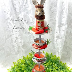 May include: A tiered stand with four levels, decorated with a bunny rabbit figurine, teacups, and carrots. The stand is made of metal and has a white and red colour scheme. The stand is surrounded by green foliage.