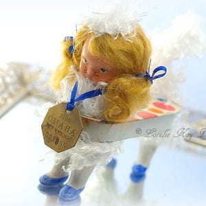 Puede incluir: Una muñeca de porcelana blanca con cabello rubio y ojos azules que lleva un vestido blanco con detalles de cinta azul. La muñeca está sentada en un pequeño carro blanco y azul con un diseño pintado de colores. La muñeca tiene una etiqueta dorada unida a su vestido que dice "2000 OMAHA PET LICENSE 00095".