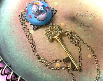 May include: A gold-coloured key pendant necklace with a blue orb pendant featuring an Alice in Wonderland design. The necklace has a delicate chain and the text "Lorelie Kay Designs".