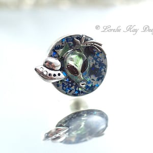 Puede incluir: Un charm de plata con un fondo de purpurina azul y verde, una cara de extraterrestre verde y una nave espacial plateada. El charm está diseñado para un brazalete de charms.