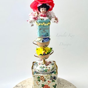 Puede incluir: Una torre de tazas de té caprichosa con una muñeca de porcelana que lleva una corona de flores rojas, una lata de té azul y blanca que dice "Traditional Afternoon Tea" y una caja con estampado floral. La torre está sobre una superficie de mármol.