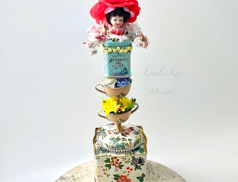 May include: A whimsical teacup tower with a porcelain doll wearing a red flower crown, a blue and white tea tin that says "Traditional Afternoon Tea", and a floral patterned box. The tower is on a marble surface.